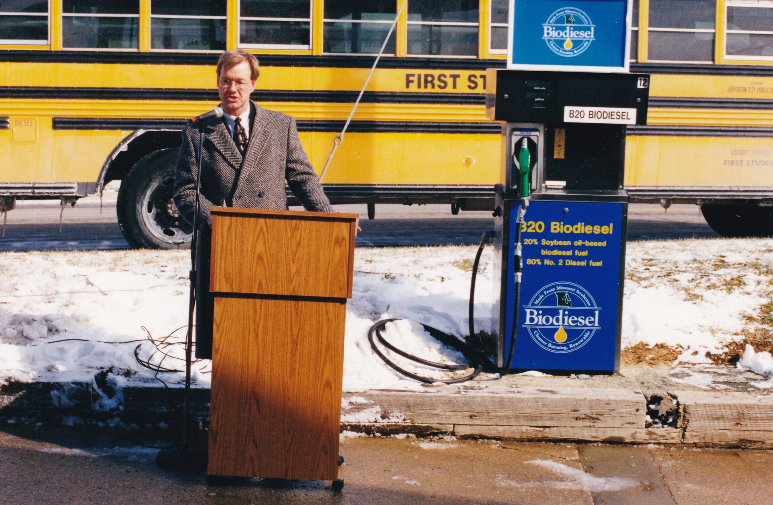 Man giving a speech in front of a biodiesel pump and a school bus